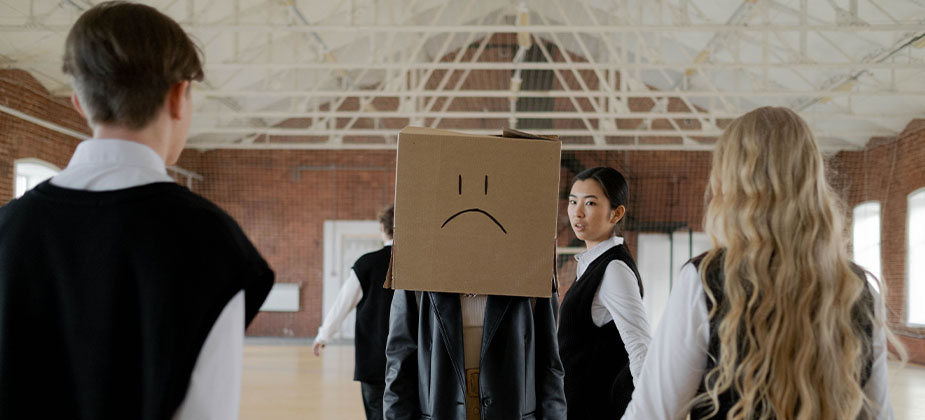 Een persoon met een doos over het hoofd met een sippe smiley, er omheen staan andere mensen die deze persoon lijken te pesten